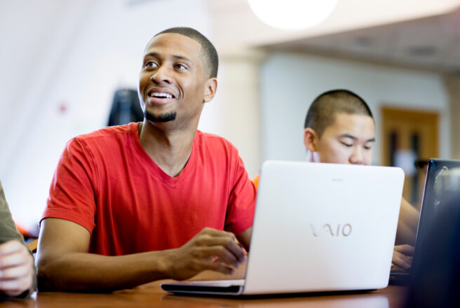 male student in red shirt smiling and using latptop